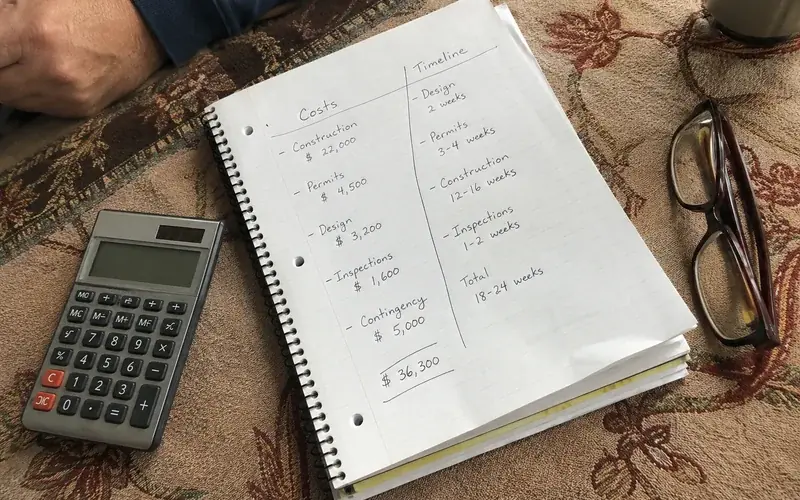 Handwritten two-column comparison of costs and timelines on a notepad beside a calculator