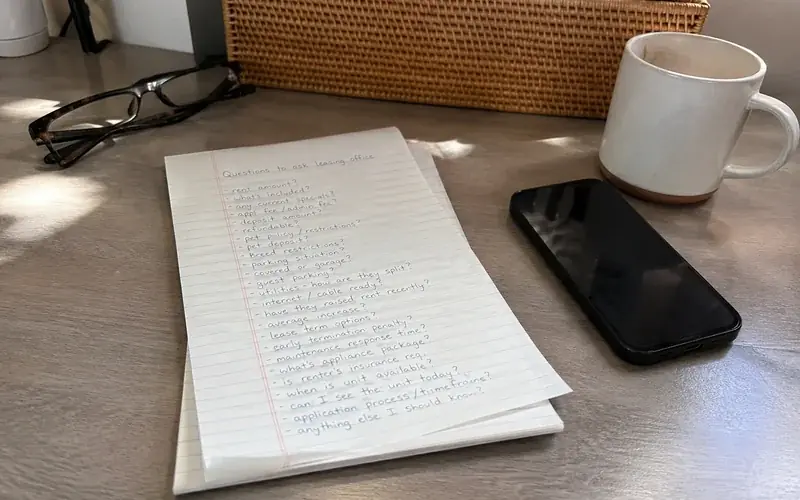 Notepad with handwritten questions to ask a leasing office beside a phone and a coffee cup
