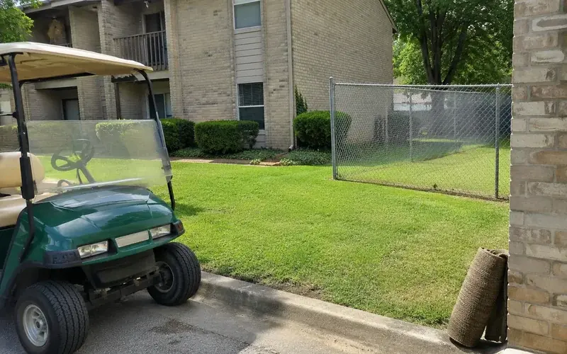 Older garden-style apartment exterior with Bermuda grass and a chain-link fence line