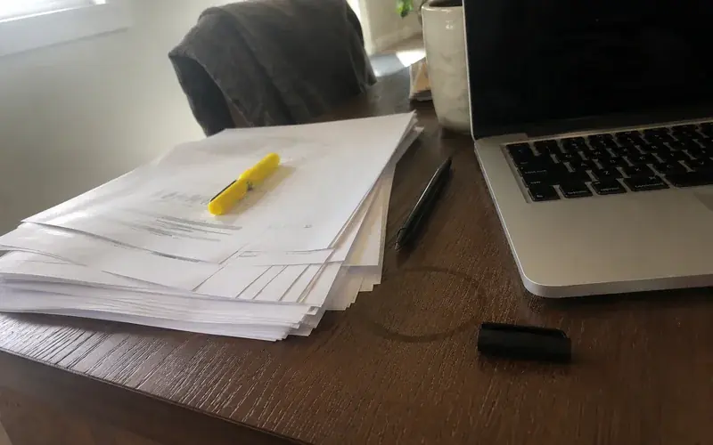 Stack of printed records and a highlighter on a home office desk beside a laptop